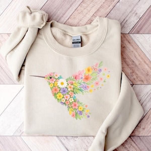 May include: A beige sweatshirt with a floral hummingbird design. The hummingbird is made up of colourful flowers in pink, yellow, purple, and white. The sweatshirt is folded on a wooden surface.