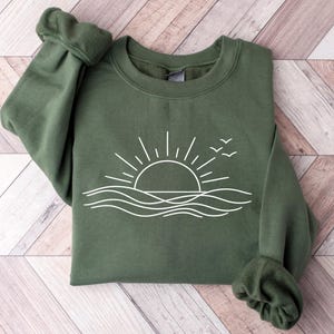 May include: Olive green sweatshirt with a white graphic of a sun setting over the ocean, with three birds flying above. The sweatshirt has a crew neck and long sleeves with ribbed cuffs.