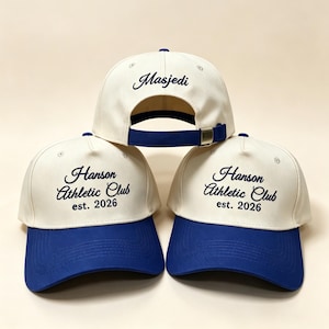 May include: Three baseball caps with a cream crown and a royal blue peak. The caps feature embroidered text: "Masjedi" on one and "Hanson Athletic Club est. 2026" on the other two. The caps have adjustable straps.