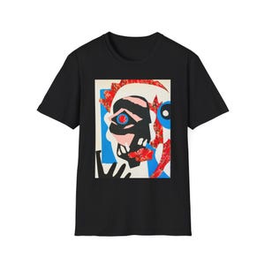 May include: Black t-shirt featuring a colorful abstract face design. The artwork includes a black face with a red eye, pink mouth, and blue and red accents. The design is centered on the front of the shirt.