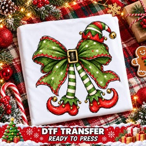 May include: A white t-shirt featuring a graphic illustration of a large green bow with red trim and a gold buckle, positioned above two striped green and white elf legs wearing curled red elf shoes with small gold bells attached.