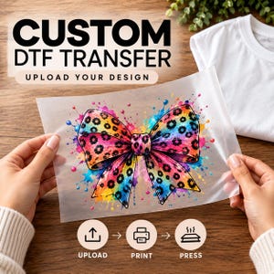 May include: A custom DTF transfer sheet featuring a colorful leopard print bow graphic with paint splatters. The text reads CUSTOM DTF TRANSFER, UPLOAD YOUR DESIGN, UPLOAD, PRINT, and PRESS. The design is held against a white t-shirt background.
