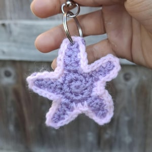 May include: A handmade crochet starfish keyring in shades of lavender and light pink. The five-pointed star is attached to a silver-coloured keyring. The crochet work features a detailed, textured pattern.