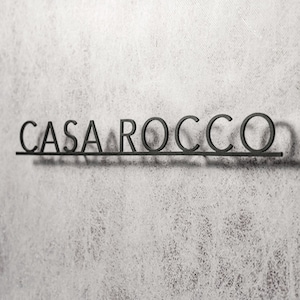 May include: Black metal sign that reads "CASA ROCCO" in a sans-serif font. The letters are connected by a horizontal bar, casting a shadow on the textured, light gray wall. The sign is a decorative element.