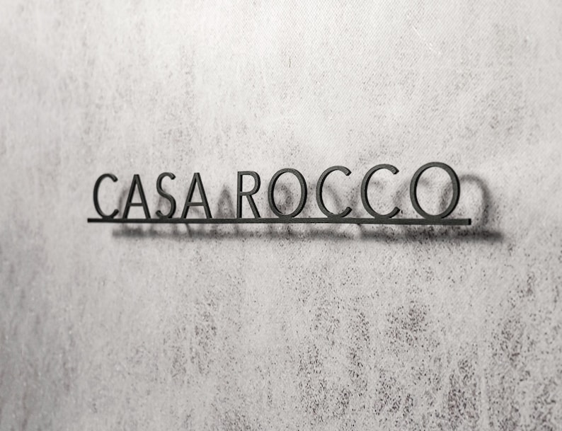May include: Black metal sign that reads "CASA ROCCO" in a sans-serif font. The letters are connected by a horizontal bar, casting a shadow on the textured, light gray wall. The sign is a decorative element.