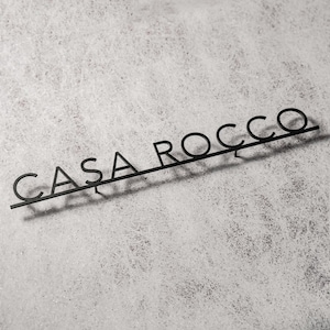 May include: Black metal sign with the text "CASA ROCCO" in a modern font. The sign is mounted on a light gray, textured surface, casting a shadow. The letters are connected by a thin bar.