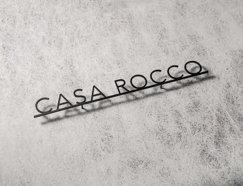 May include: Black metal sign with the text "CASA ROCCO" in a modern font. The sign is mounted on a light gray, textured surface, casting a shadow. The letters are connected by a thin bar.
