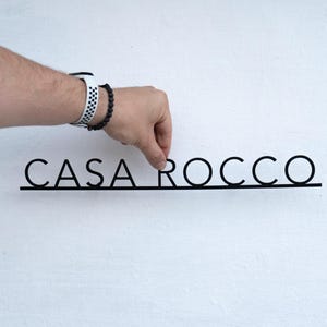 May include: Black metal sign with the text "CASA ROCCO" in a minimalist font. The sign is being held against a white wall. The letters are connected by a thin line.