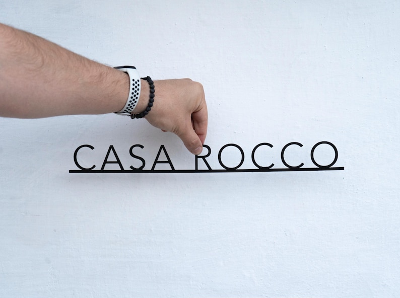 May include: Black metal sign with the text "CASA ROCCO" in a minimalist font. The sign is being held against a white wall. The letters are connected by a thin line.