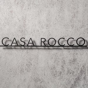 May include: Black metal sign with the text "CASA ROCCO" in a sans-serif font. The letters are connected by a horizontal line. The sign is mounted on a textured, light gray wall.