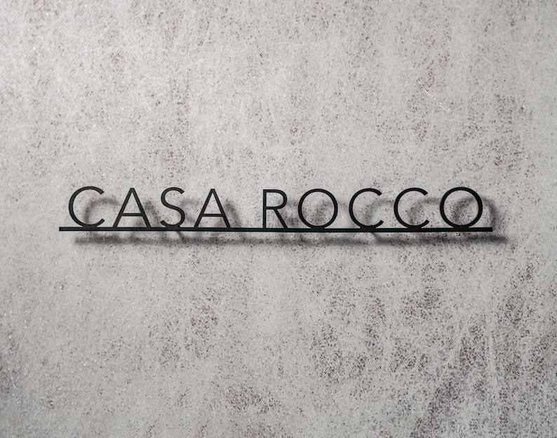 May include: Black metal sign with the text "CASA ROCCO" in a sans-serif font. The letters are connected by a horizontal line. The sign is mounted on a textured, light gray wall.
