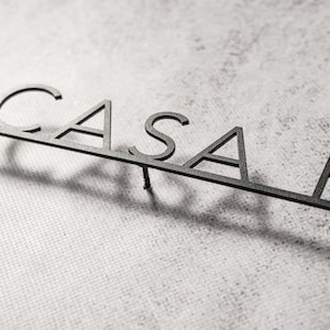 May include: A black metal sign that spells out "CASAR" in a modern, minimalist font. The letters are connected by a thin bar, and the sign casts a shadow on a textured, light-colored surface.