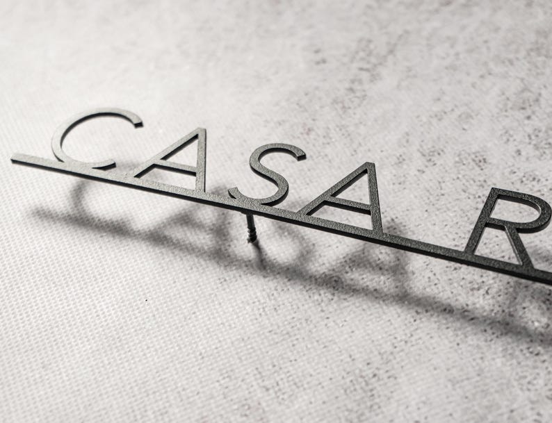 May include: A black metal sign that spells out "CASAR" in a modern, minimalist font. The letters are connected by a thin bar, and the sign casts a shadow on a textured, light-colored surface.