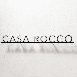 May include: Black metal sign that reads "CASA ROCCO" in a minimalist font. The letters are connected by a thin horizontal line, casting a shadow on the white textured wall. The sign is a decorative element.