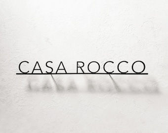 Puede incluir: Letrero de metal negro que dice "CASA ROCCO" en una fuente minimalista. Las letras están conectadas por una fina línea horizontal, proyectando una sombra sobre la pared blanca texturizada. Un elemento decorativo.