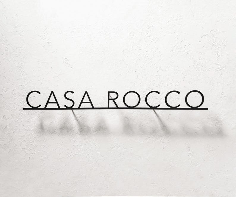 May include: Black metal sign that reads "CASA ROCCO" in a minimalist font. The letters are connected by a thin horizontal line, casting a shadow on the white textured wall. The sign is a decorative element.