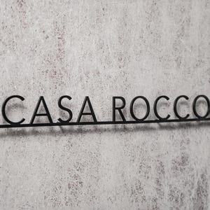 May include: Black metal sign with the text "CASA ROCCO" in a sans-serif font. The letters are connected by a thin horizontal line. The sign is mounted on a textured, light gray surface.