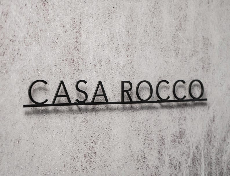 May include: Black metal sign with the text "CASA ROCCO" in a sans-serif font. The letters are connected by a thin horizontal line. The sign is mounted on a textured, light gray surface.