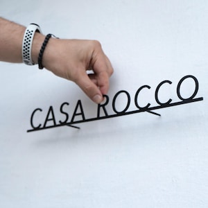 May include: Black metal sign that reads "CASA ROCCO" in a modern font. The sign is being held against a white wall. The sign is likely for home decor.