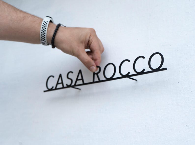 May include: Black metal sign that reads "CASA ROCCO" in a modern font. The sign is being held against a white wall. The sign is likely for home decor.