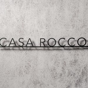 May include: Black metal sign with the words "CASA ROCCO" in a minimalist font. The letters are connected by a horizontal line, casting a shadow on the textured, light gray background. The sign is designed for indoor or outdoor use.