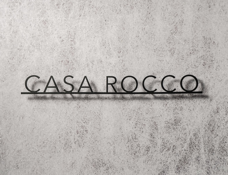 May include: Black metal sign with the words "CASA ROCCO" in a minimalist font. The letters are connected by a horizontal line, casting a shadow on the textured, light gray background. The sign is designed for indoor or outdoor use.