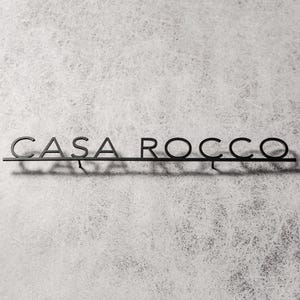 May include: A black metal sign that reads "CASA ROCCO" in a minimalist font. The sign is mounted on a thin horizontal bar, casting a shadow against a textured, light gray background. The letters are connected.