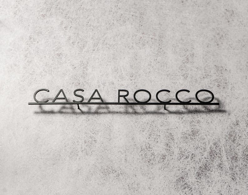 May include: A black metal sign that reads "CASA ROCCO" in a minimalist font. The sign is mounted on a thin horizontal bar, casting a shadow against a textured, light gray background. The letters are connected.