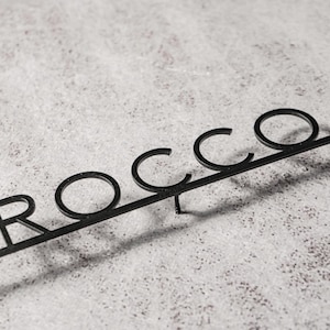 May include: Black metal sign that spells out the name "ROCCO" in a stylized font. The letters are connected by a horizontal bar, with a small stake at the bottom for insertion. The sign is set against a textured, light-colored background.