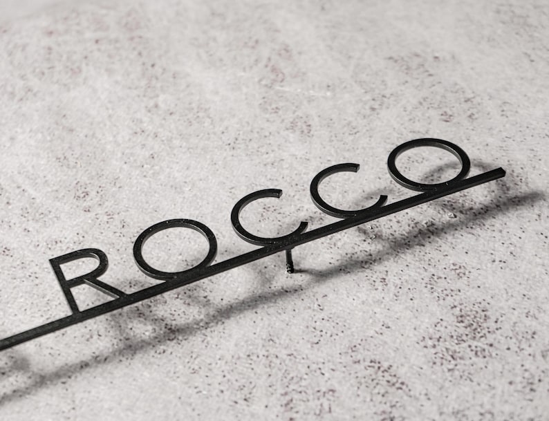 May include: Black metal sign that spells out the name "ROCCO" in a stylized font. The letters are connected by a horizontal bar, with a small stake at the bottom for insertion. The sign is set against a textured, light-colored background.