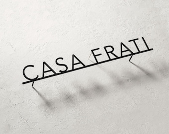 Puede incluir: Letrero de pared de metal negro con el texto "CASA FRATI" en una fuente moderna. El letrero está montado en una pared blanca texturizada, proyectando una sombra. Es un elemento decorativo para la decoración del hogar.