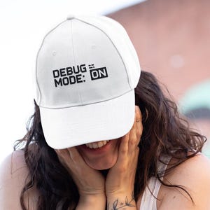 May include: White baseball cap with the text "DEBUG MODE: ON" in black. The cap is worn by a person. The person is wearing a white vest top and black jeans. The background is blurred, with a pink and grey wall.