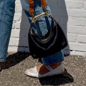 May include: A black, curved handbag with a gold zip and a furry texture. It has a brown leather strap. The person is wearing blue jeans and white ballet flats. The bag's strap is brown leather.