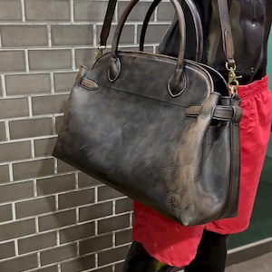 May include: A distressed leather handbag with a top handle and a detachable shoulder strap. The bag is dark brown with lighter brown accents and gold-colored hardware. The bag is displayed on a mannequin.