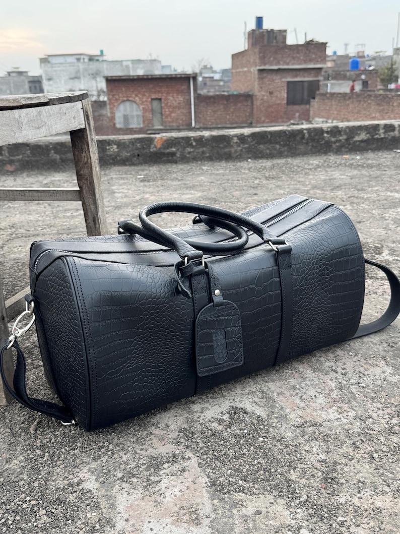 Op de afbeelding: Zwarte duffeltas met een textuur die op krokodillenleer lijkt. De cilindervormige tas heeft een ritssluiting aan de bovenkant, twee handvatten en een afneembare schouderriem. Een klein label is bevestigd.
