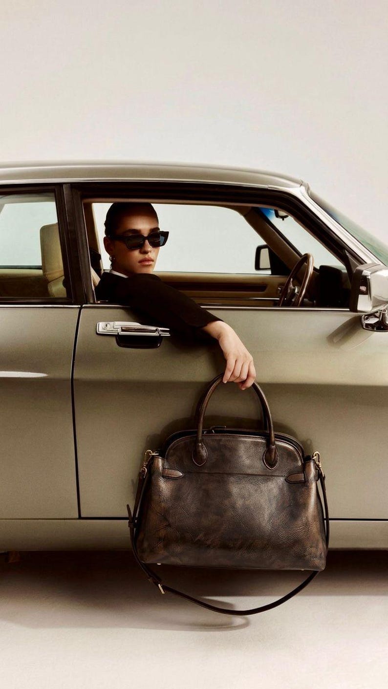 May include: A dark brown leather handbag with a top handle and shoulder strap. The bag is held by a person in a car. The car is a light green colour.