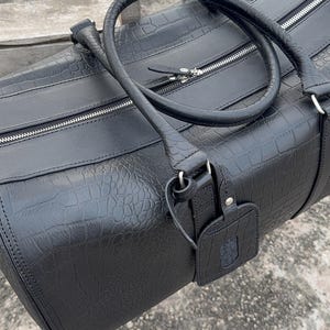 Op de afbeelding: Zwarte duffeltas met een textuur die op krokodillenleer lijkt. Voorzien van een handvat aan de bovenkant, ritssluiting en een bagagelabel. De tas is gemaakt van duurzaam materiaal, geschikt voor reizen. Ongeveer 60 cm lang.