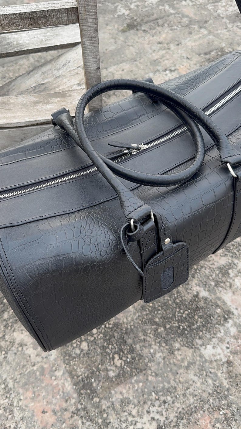 Op de afbeelding: Zwarte duffeltas met een textuur die op krokodillenleer lijkt. Voorzien van een handvat aan de bovenkant, ritssluiting en een bagagelabel. De tas is gemaakt van duurzaam materiaal, geschikt voor reizen. Ongeveer 60 cm lang.