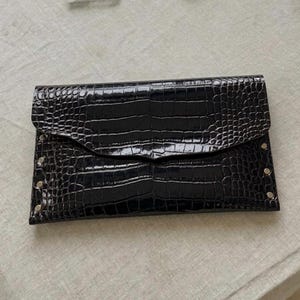 May include: A black clutch with a crocodile skin texture. The clutch has a flap closure and is secured with small silver rivets. The accessory is rectangular and appears to be made of leather or a similar material.