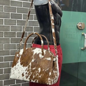 Op de afbeelding: Een bruin-witte koeienhuid handtas met een handvat aan de bovenkant en een afneembare schouderriem. De tas wordt getoond op een mannequin met een rode korte broek. De tas heeft een gestructureerde vorm en zilverkleurige hardware.