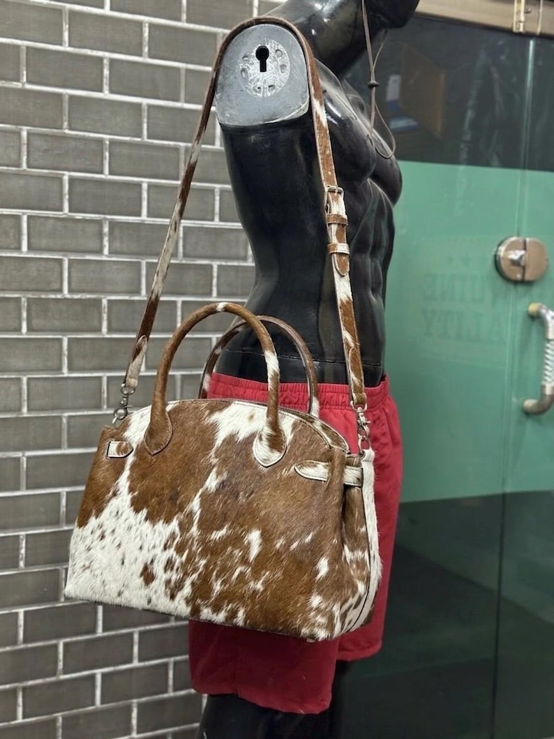 Op de afbeelding: Een bruin-witte koeienhuid handtas met een handvat aan de bovenkant en een afneembare schouderriem. De tas wordt getoond op een mannequin met een rode korte broek. De tas heeft een gestructureerde vorm en zilverkleurige hardware.
