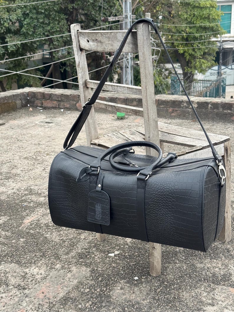 Op de afbeelding: Een zwarte duffeltas met een textuur die op krokodillenleer lijkt. De tas heeft een handvat aan de bovenkant, een schouderriem en een bagagelabel. De tas hangt aan een houten stoel.