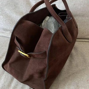May include: A brown suede tote bag with two handles. The bag has a rectangular shape with a gold-colored buckle on the side. The interior of the bag is visible, and it appears to be empty.