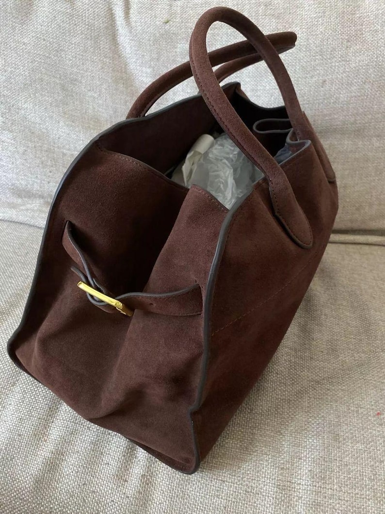May include: A brown suede tote bag with two handles. The bag has a rectangular shape with a gold-colored buckle on the side. The interior of the bag is visible, and it appears to be empty.