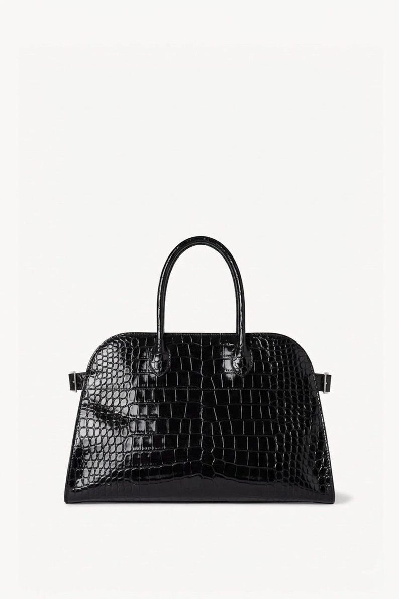 Puede incluir: Bolso de mano negro brillante con textura de piel de cocodrilo. El bolso tiene una parte superior redondeada, un asa curva y dos peque&ntilde;as hebillas en los laterales. Fondo blanco.