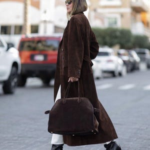 May include: A woman wearing a long brown suede coat, white pants, and black pointed-toe boots. She carries a large brown suede handbag. The coat has a belted waist and button details. The background shows a city street with cars.