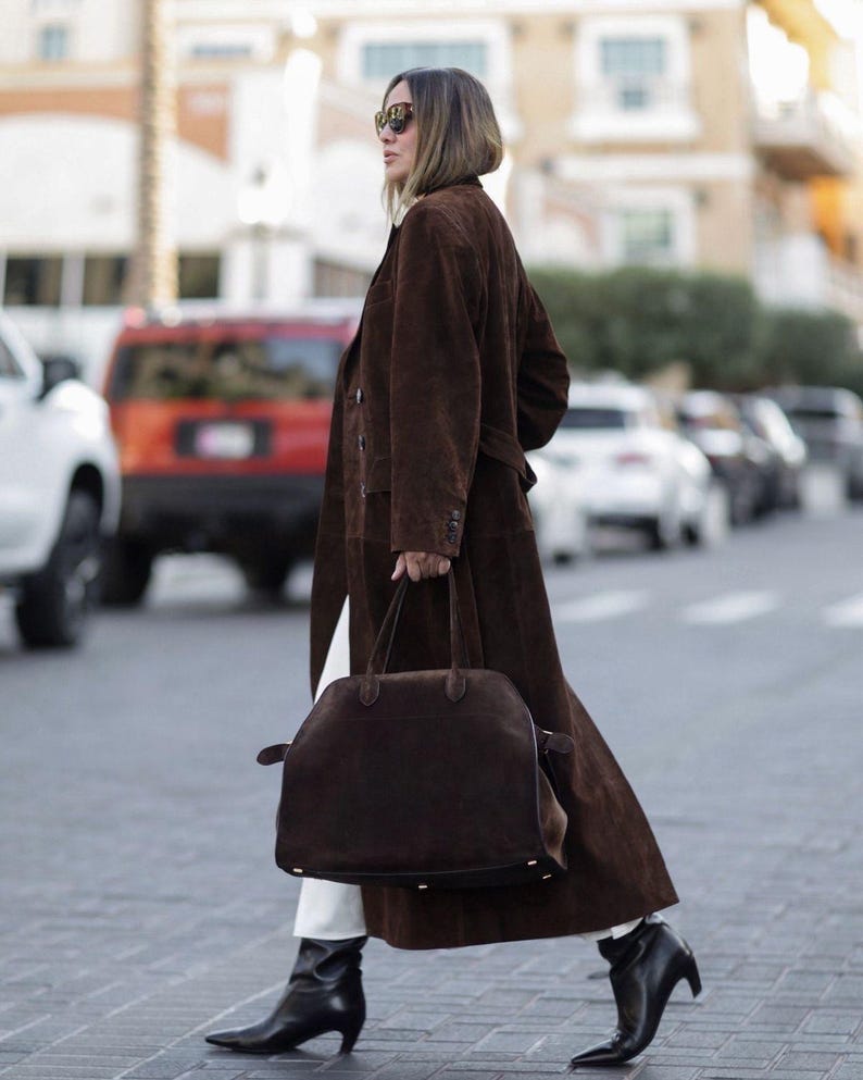 May include: A woman wearing a long brown suede coat, white pants, and black pointed-toe boots. She carries a large brown suede handbag. The coat has a belted waist and button details. The background shows a city street with cars.