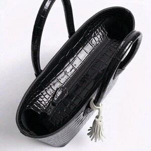 May include: Black tote bag with a glossy, crocodile-skin texture. The bag has two rounded handles and a zipper closure. A small, white tassel hangs from the side. The interior is visible.