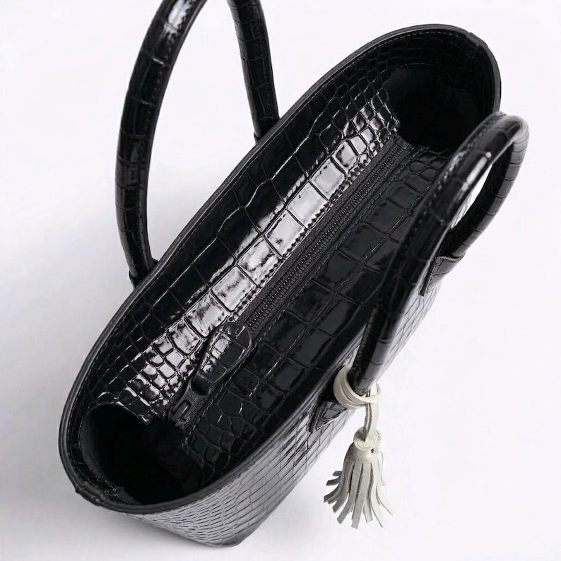 May include: Black tote bag with a glossy, crocodile-skin texture. The bag has two rounded handles and a zipper closure. A small, white tassel hangs from the side. The interior is visible.