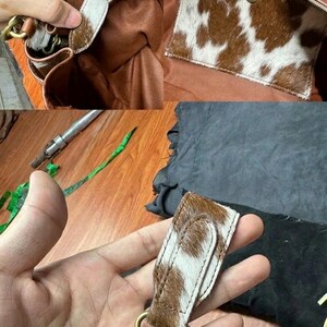 Op de afbeelding: Een bruine leren handtas met een koeienhuidpatroon. De tas heeft een ritssluiting, een binnenvak en een afneembare riem met een messing sluiting. Het koeienhuidpatroon is bruin en wit.